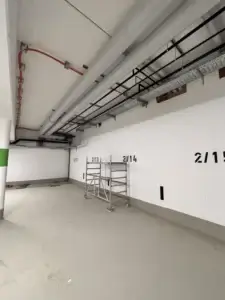 Podstropné káblové žľaby a rozvody potrubí v podzemnej garáži s označenými parkovacími miestami a lešením.