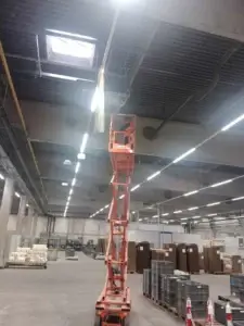 Výškové práce na inštalácii káblových trás v priemyselnej hale pomocou nožnicovej plošiny a osvetlením pod stropom.